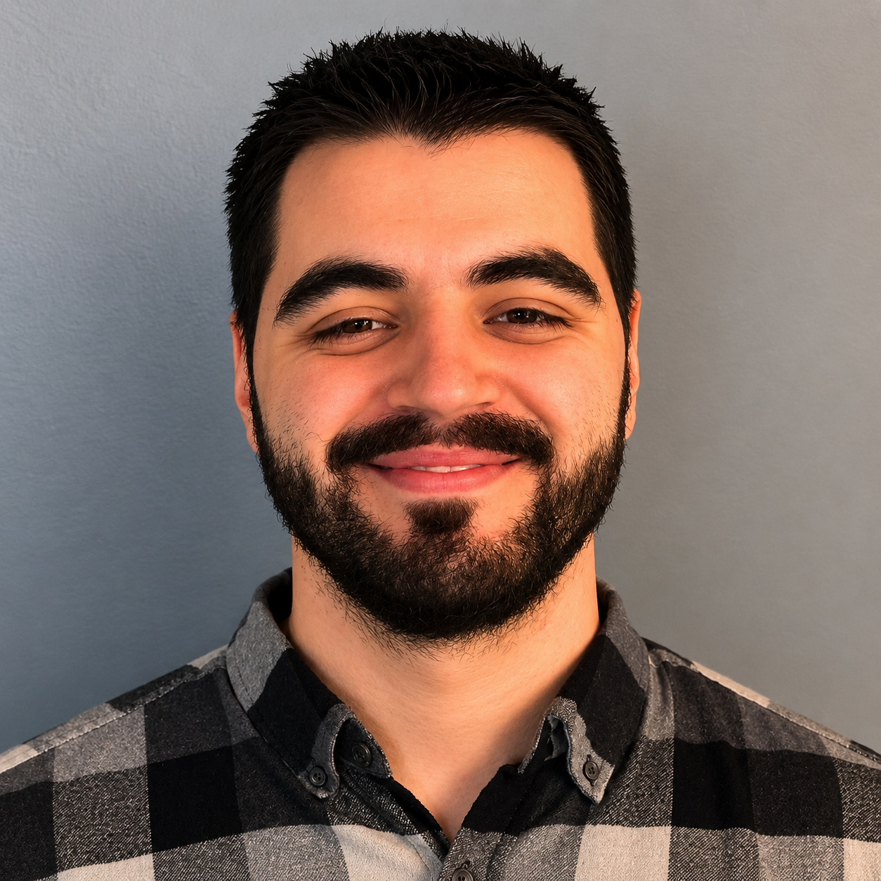 Smiling bearded man in a black-and-gray plaid shirt against a gray background