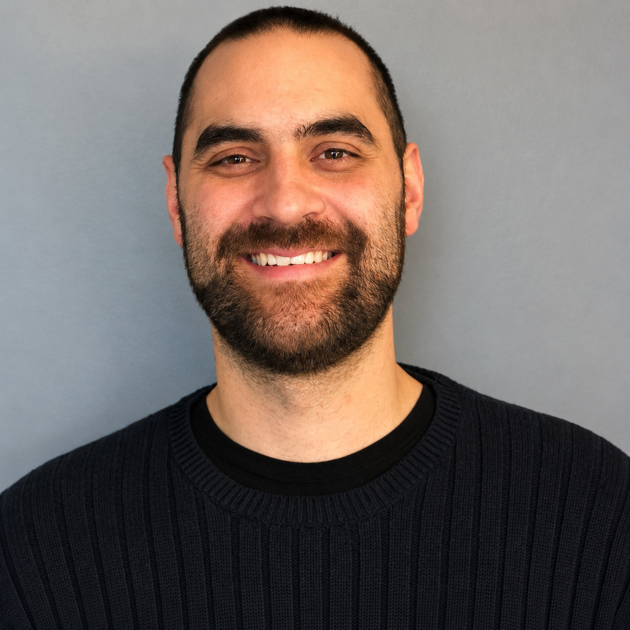 Smiling man with short dark hair and beard, wearing a black sweater against a gray background