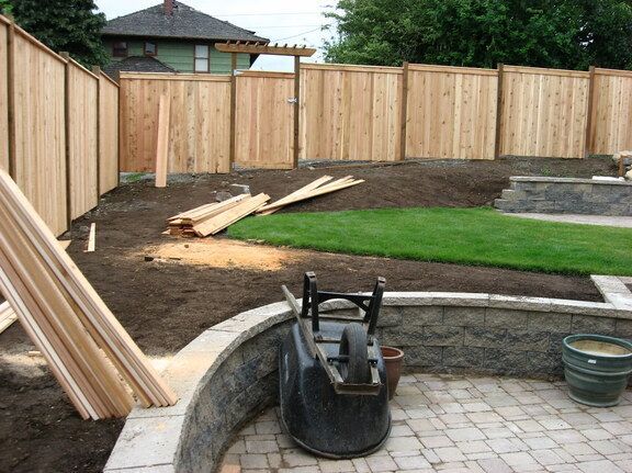Backyard under construction with wooden fence, dirt, pavers, and stacked lumber near a patio.