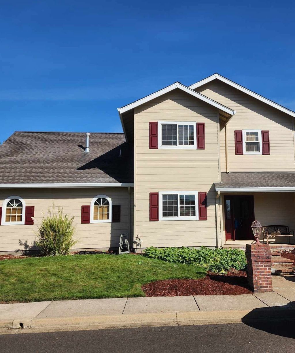 Two-story beige suburban house with red shutters, front lawn, and brick mailbox under a blue sky
