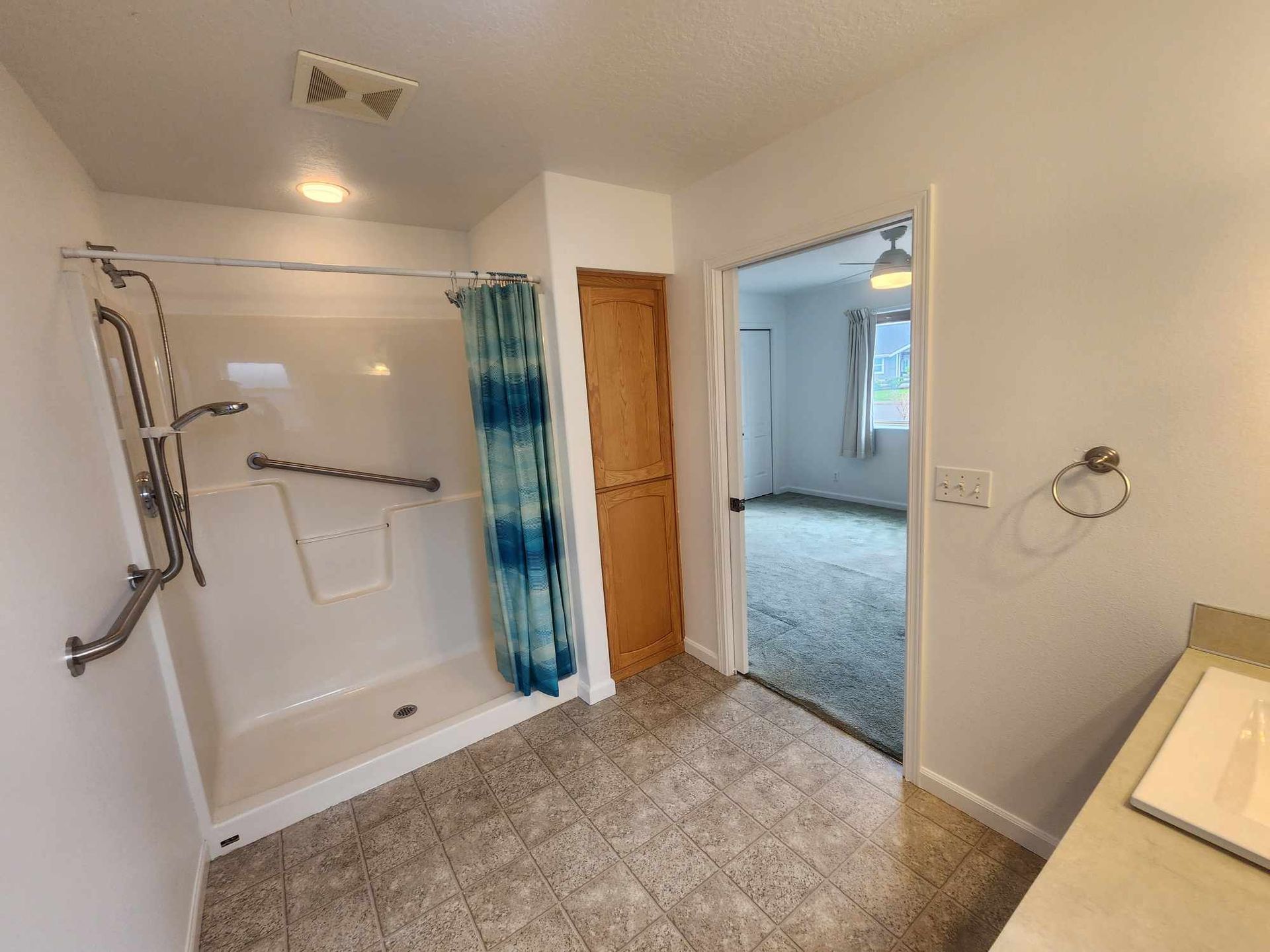 Accessible bathroom with walk-in shower, blue curtain, grab bars, and open doorway to a bedroom.