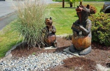 Two carved wooden bear statues in a garden setting; one holds a sign, the other sits.