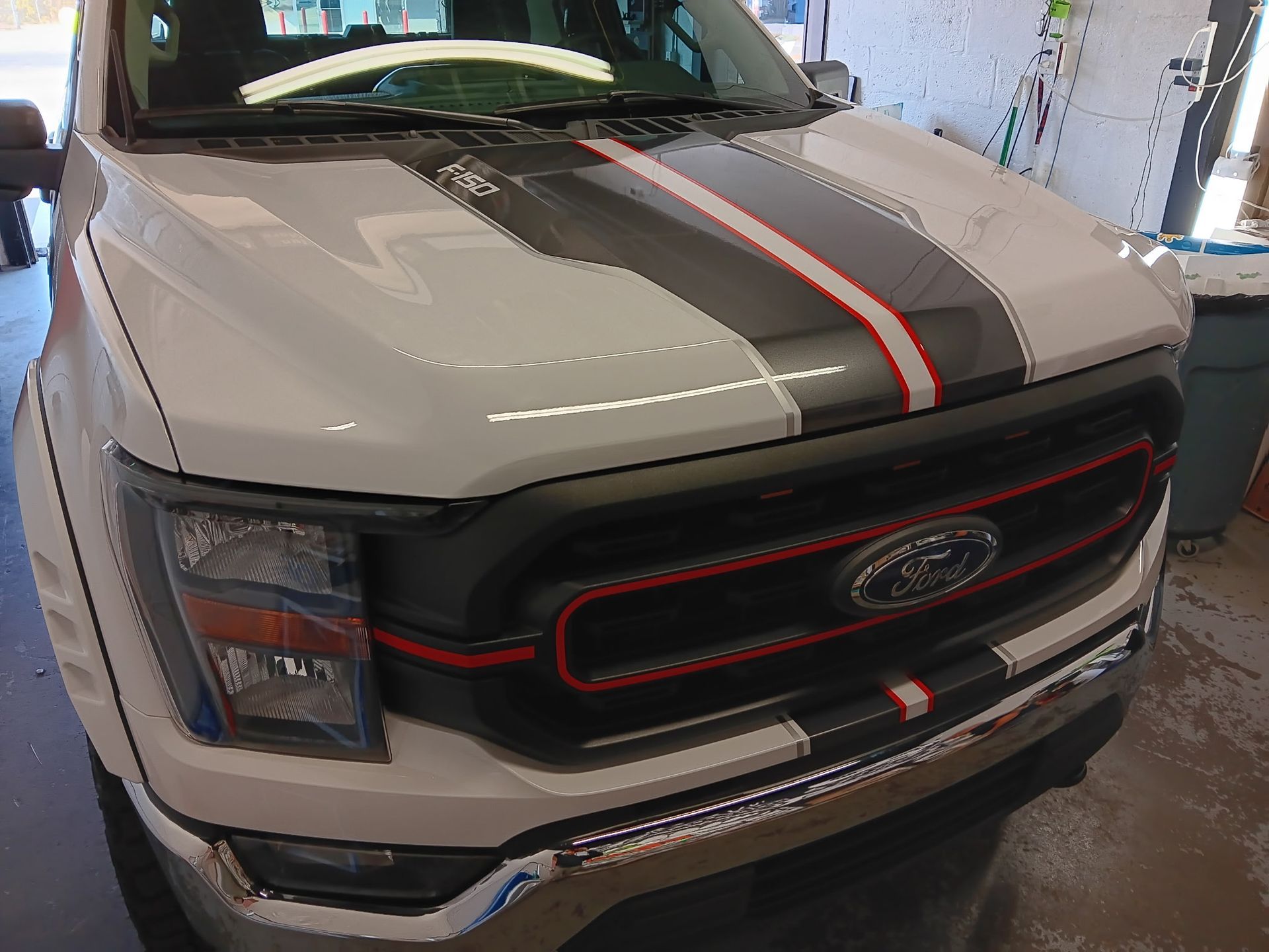 A white ford truck with a black and red stripe on the hood is parked in a garage.