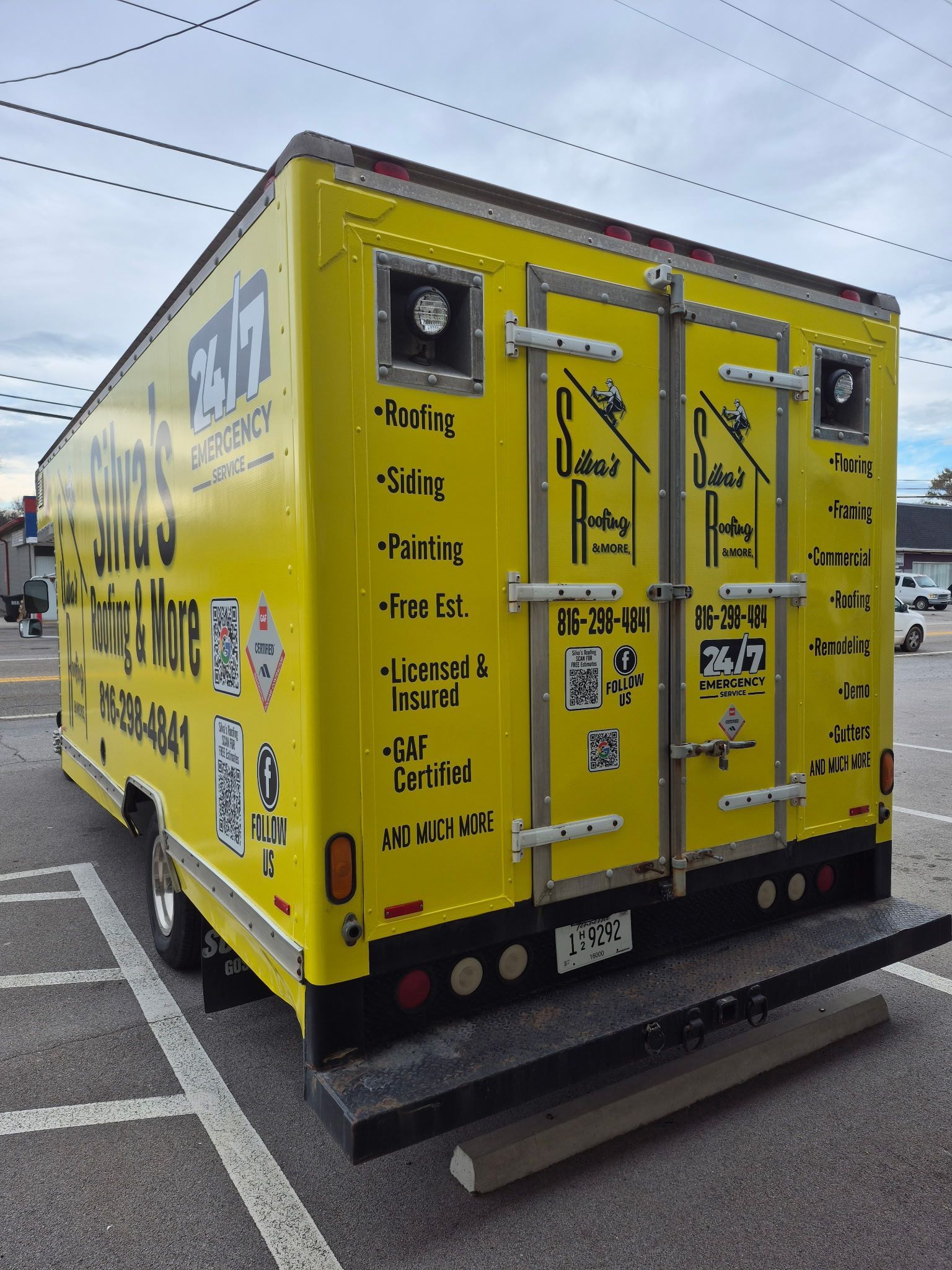 Yellow moving truck with advertisements parked on a street. Rear doors are closed, with a pull-down ramp.