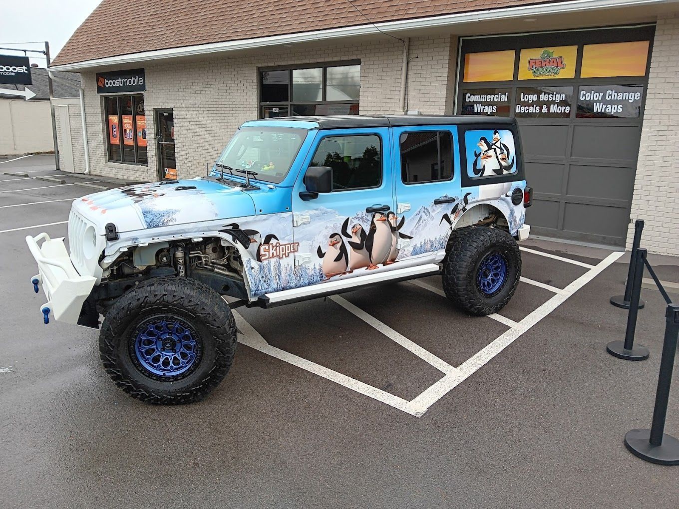 A blue and white jeep is parked in a parking lot in front of a building.