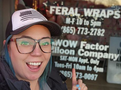 A woman wearing glasses and a hat stands in front of a window that says feral wraps