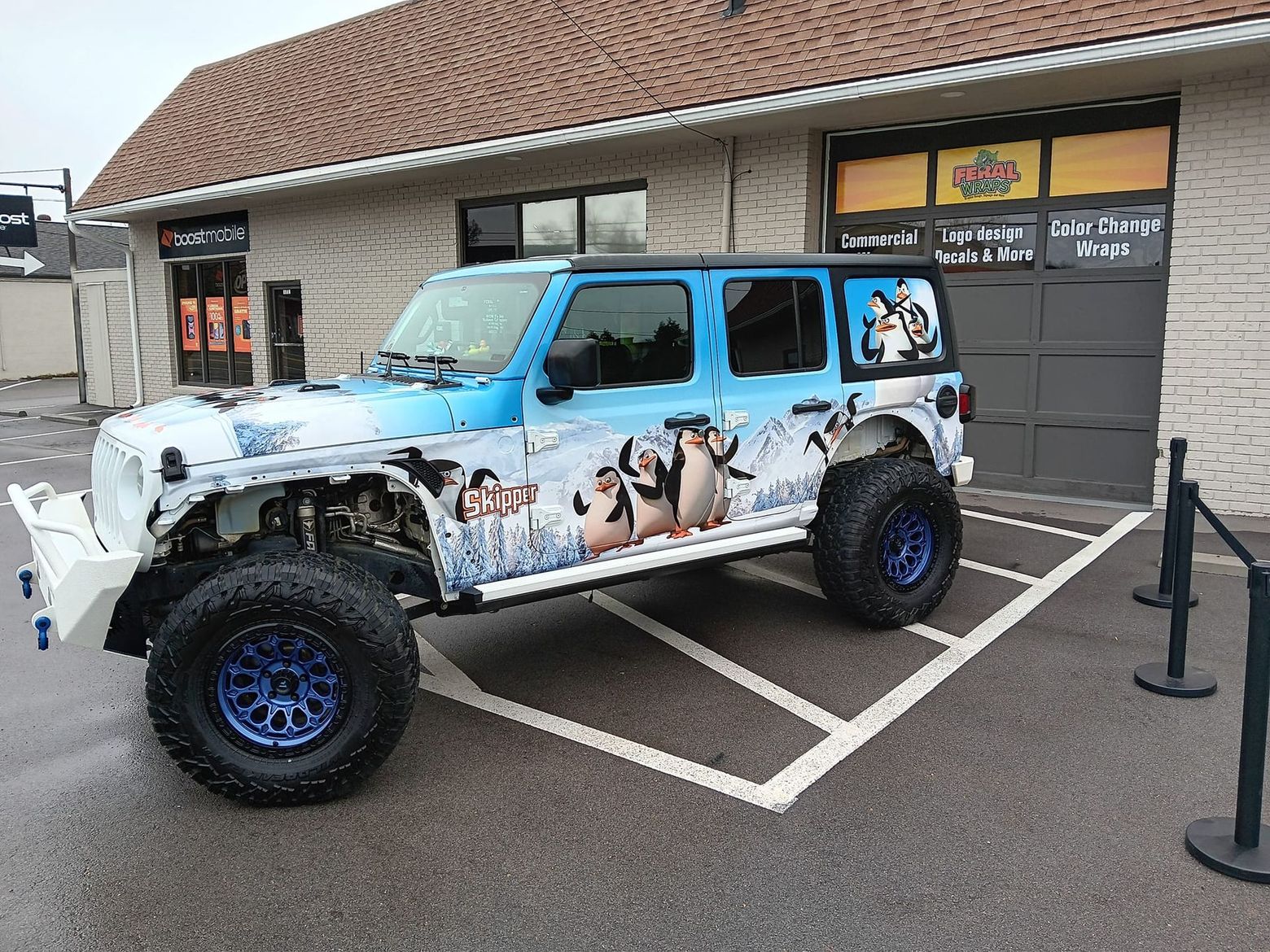 A blue and white jeep is parked in a parking lot in front of a building.