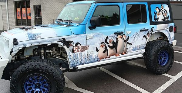 A blue and white jeep with penguins on the side is parked in a parking lot.