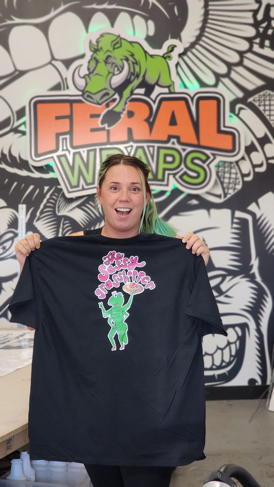 A woman is holding a black t-shirt in front of a feral wraps sign.