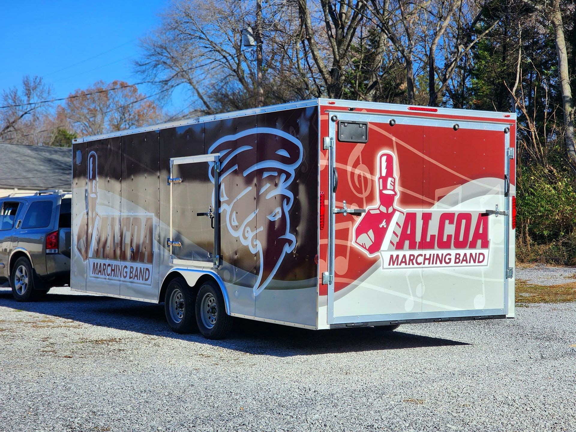 A trailer with a marching band logo on it is parked in a gravel lot.
