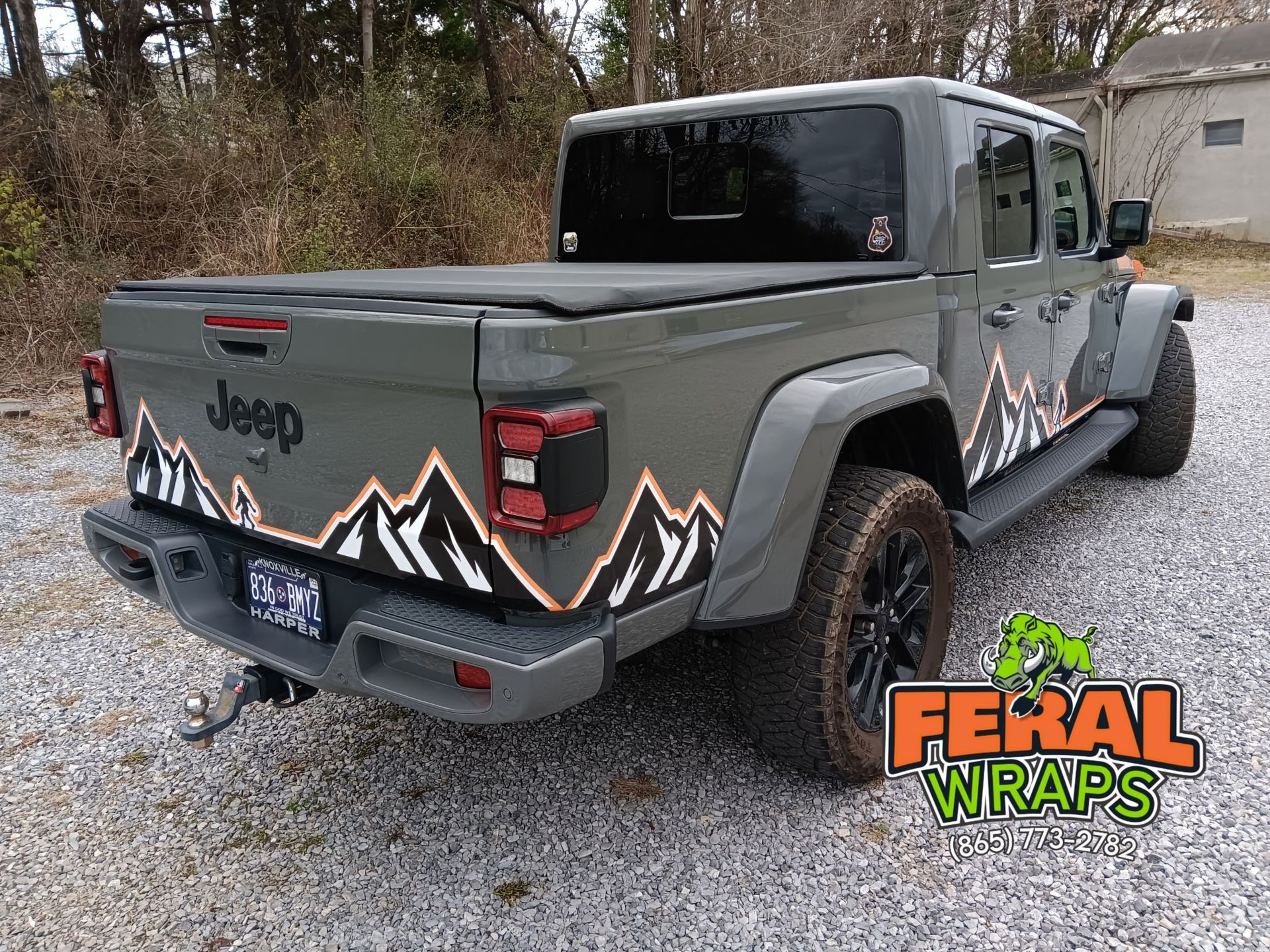 A jeep gladiator with mountains on the back is parked in a gravel lot.