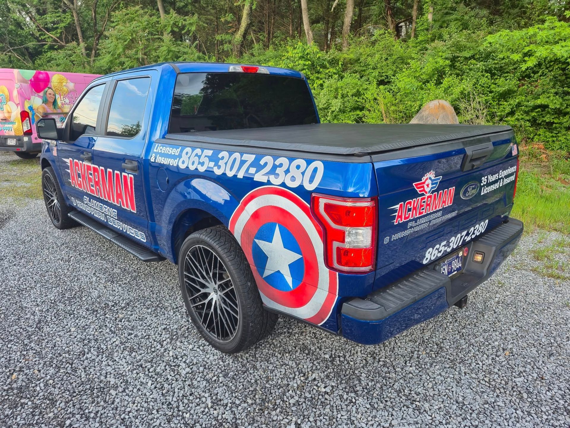 A blue truck with a captain america shield on the back is parked in a gravel lot.
