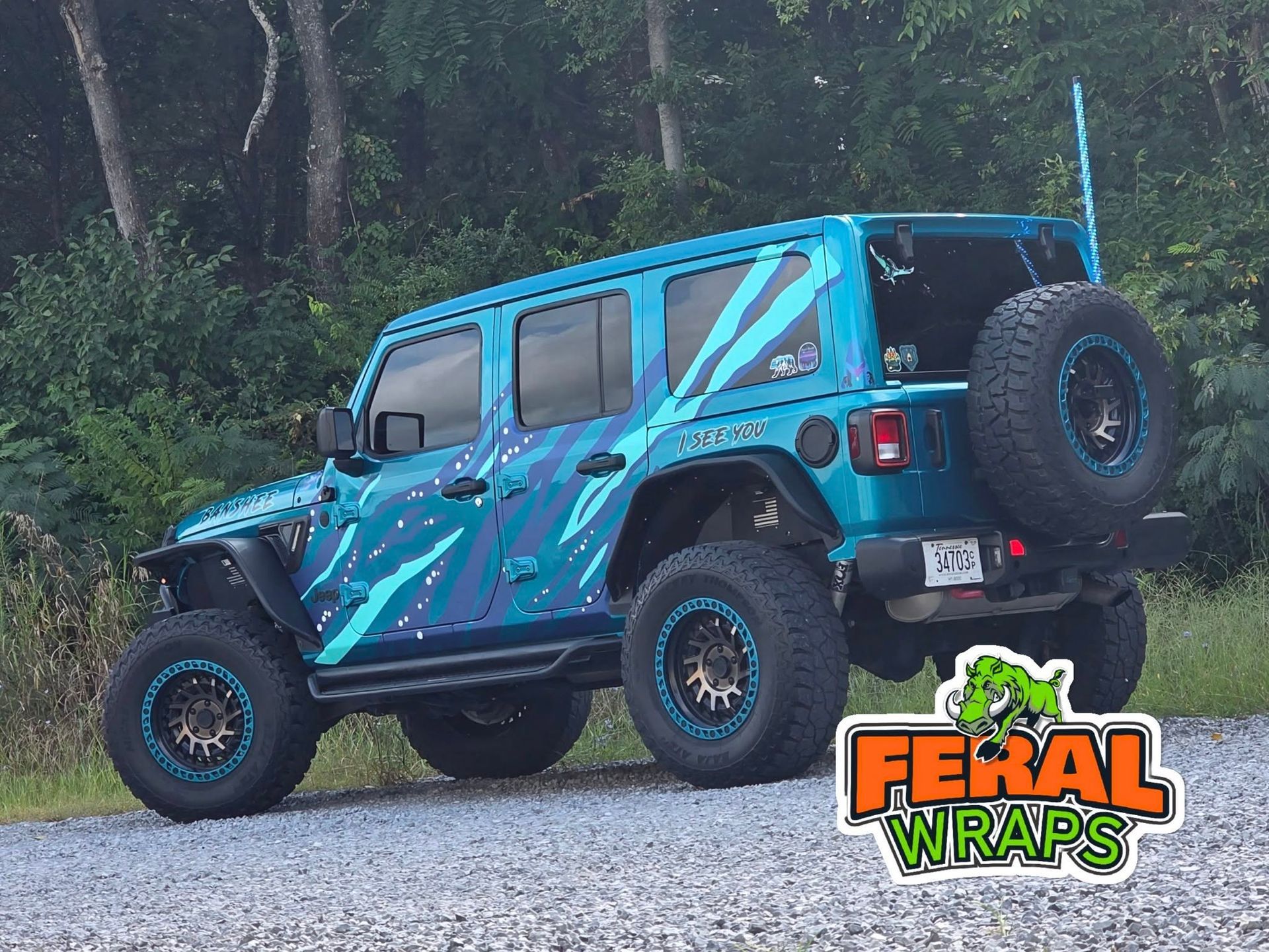 Blue Jeep with custom wrap on a gravel road, logo reads 