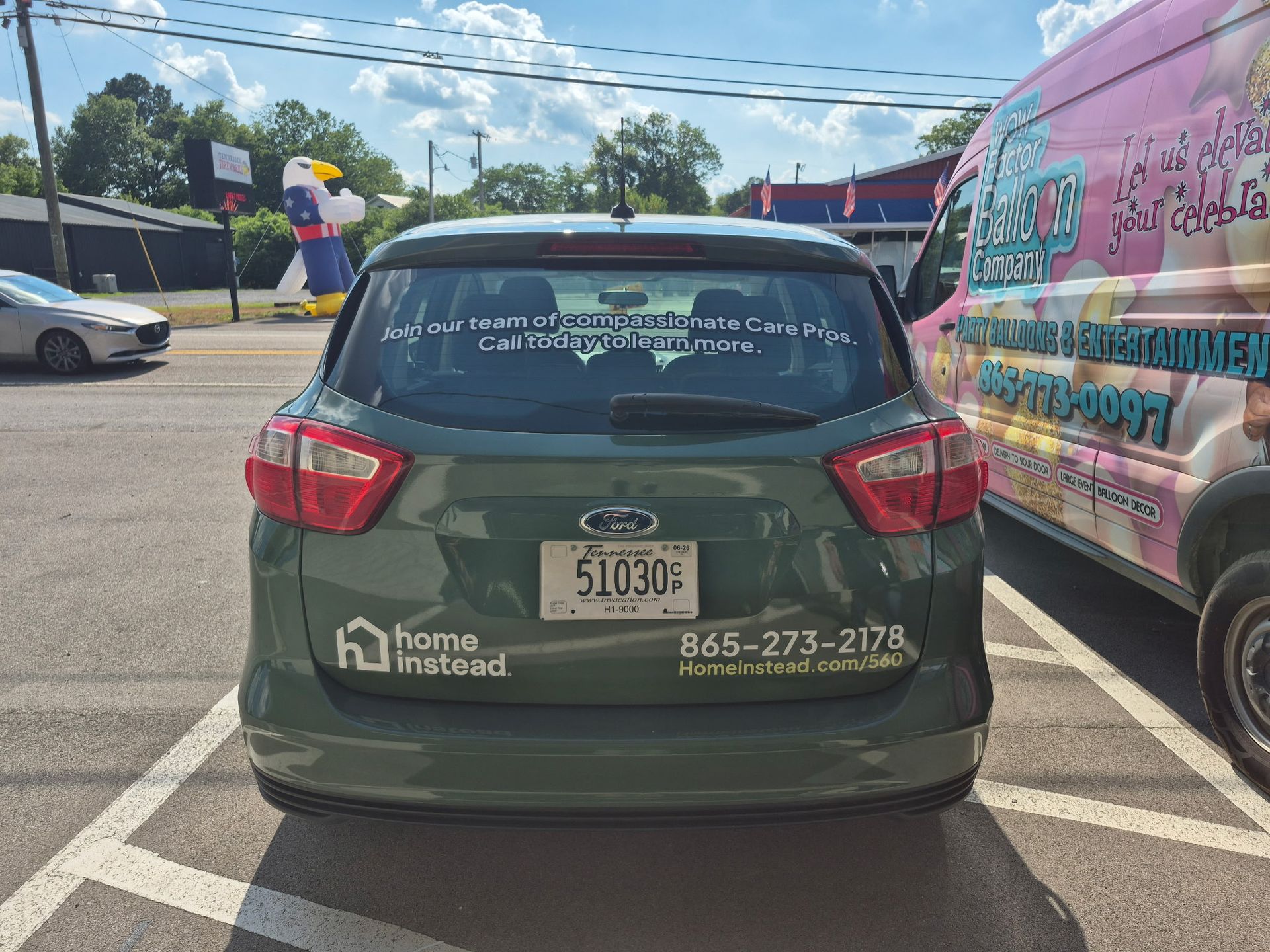 A green car is parked in a parking lot next to a pink van.