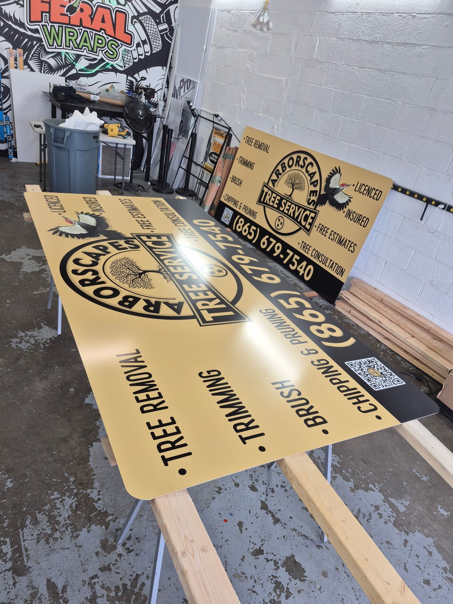 Signs for Arborist service, gold and black with contact info, sitting on a wooden stand in a workshop.