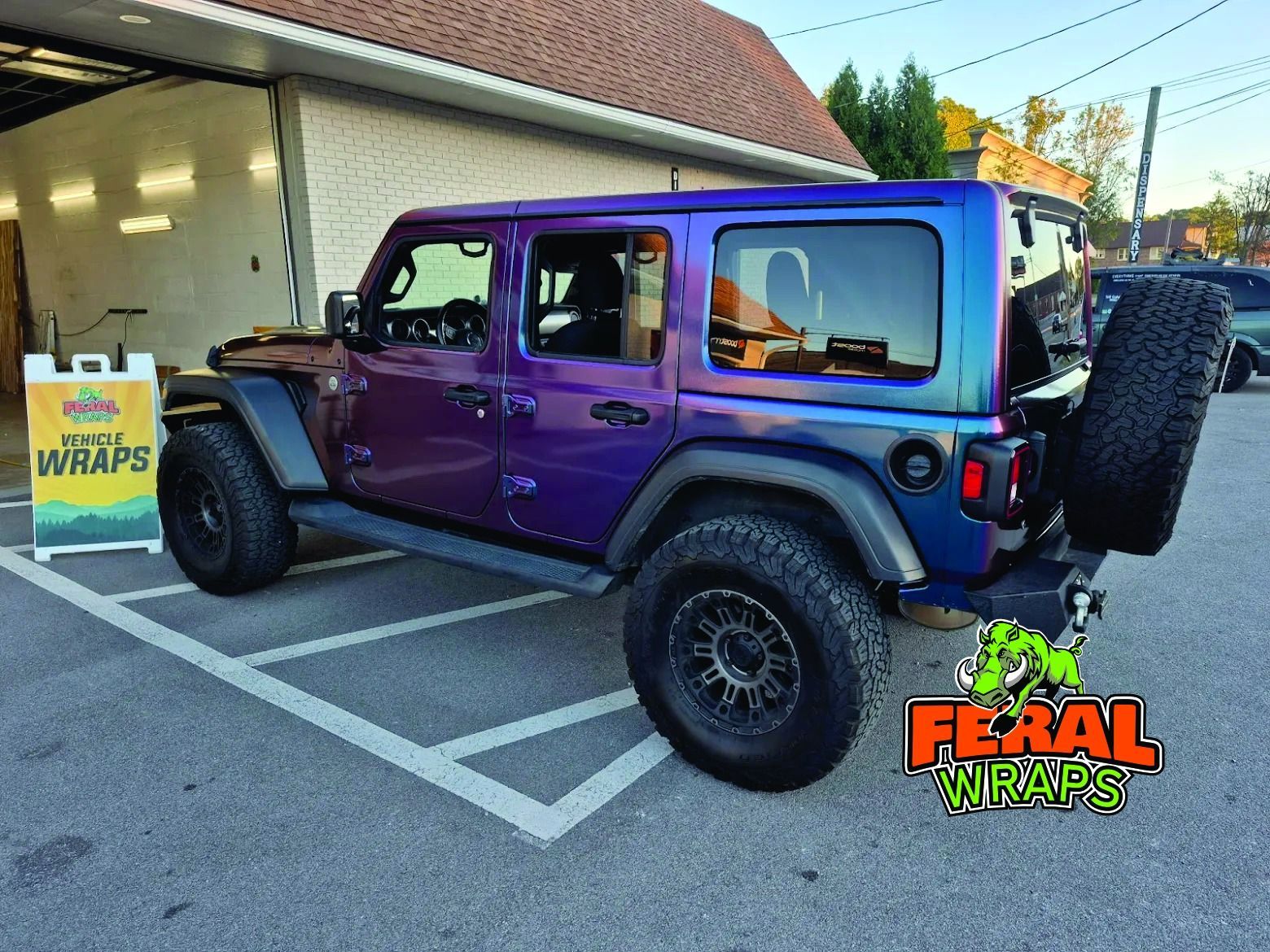 A purple and blue wrap on a Jeep Wrangler parked outside a shop. Black wheels, spare tire, and shop sign are visible.