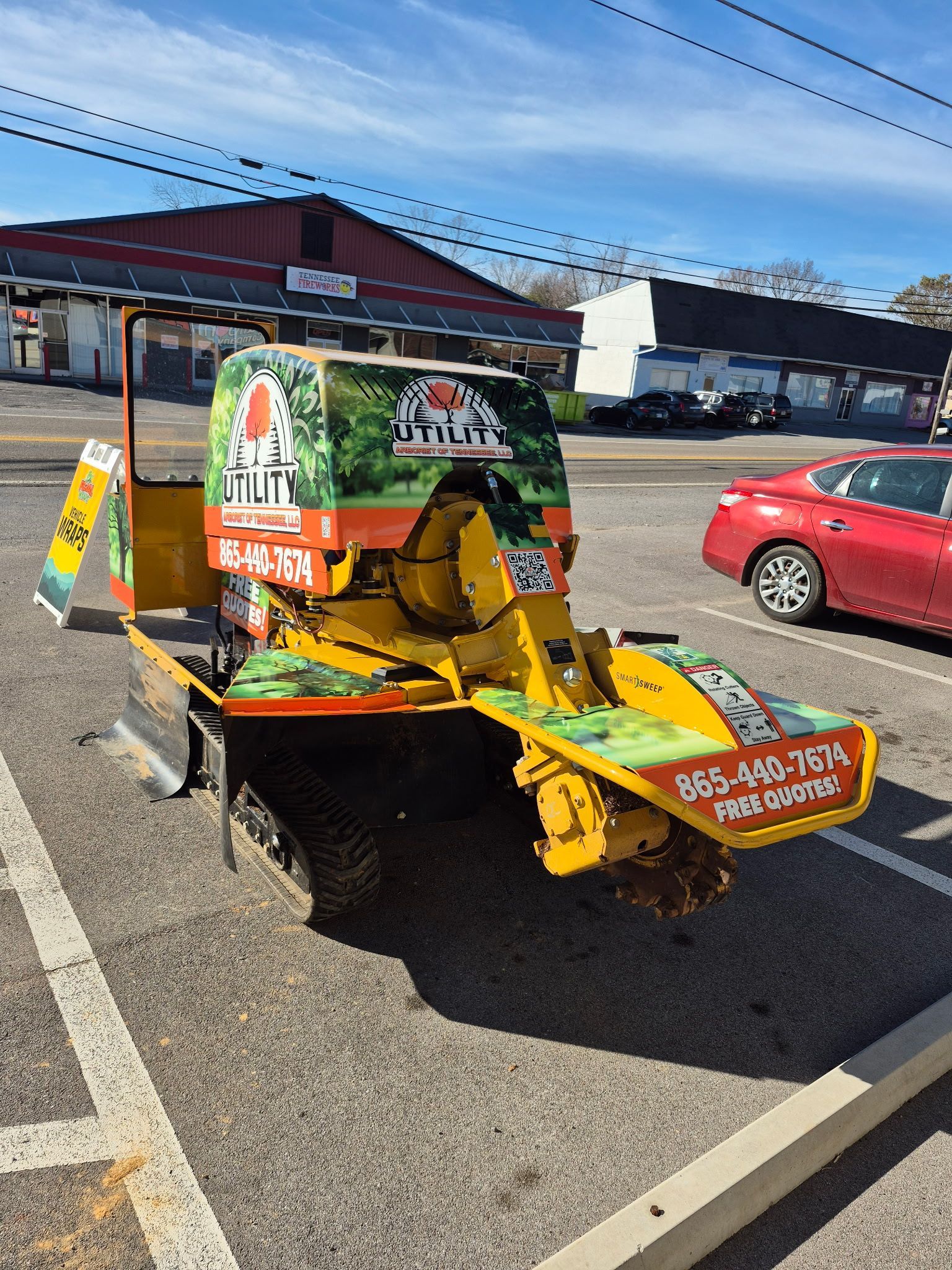 A yellow stump grinder is parked in a parking lot next to a red car.