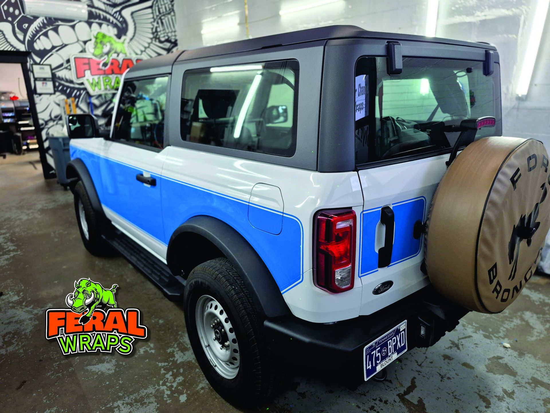Ford Bronco with blue and white wrap, black roof, and spare tire, inside a garage.