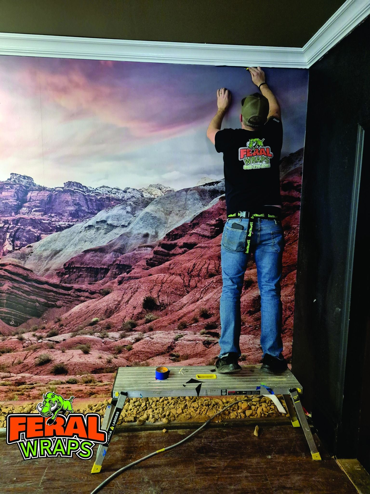 Person installing crown molding on a wall with a mountain mural, standing on a step platform.