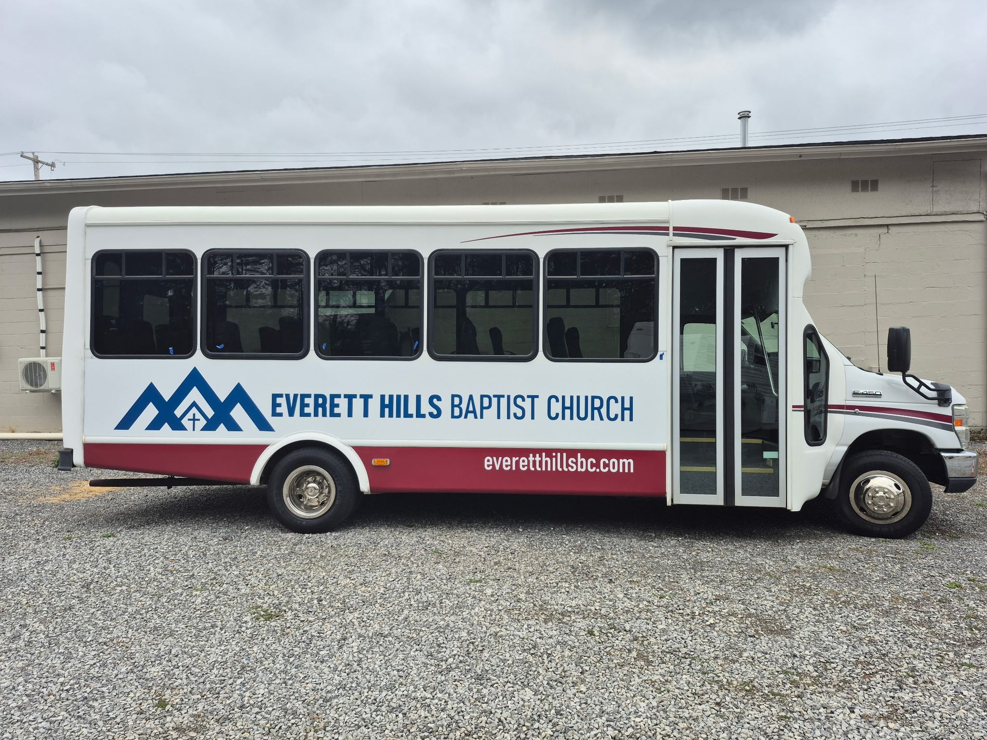 White bus with Everett Hills Baptist Church logo, parked on gravel. Red and blue accents.