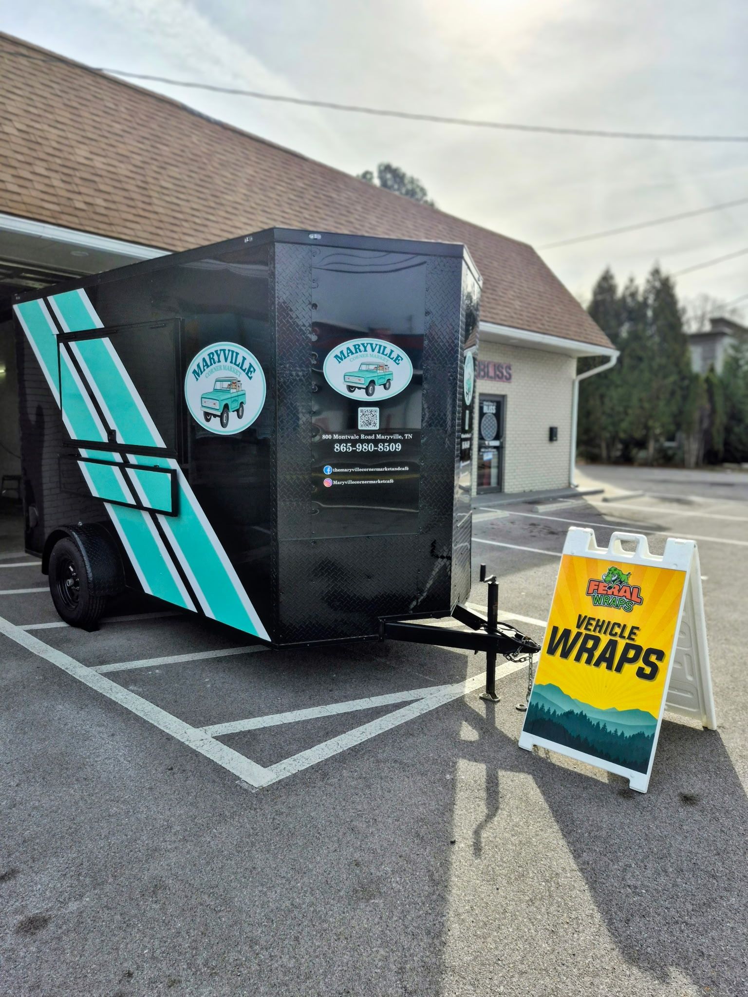 Black trailer with turquoise stripes and logo, parked in front of a building. A-frame sign says 