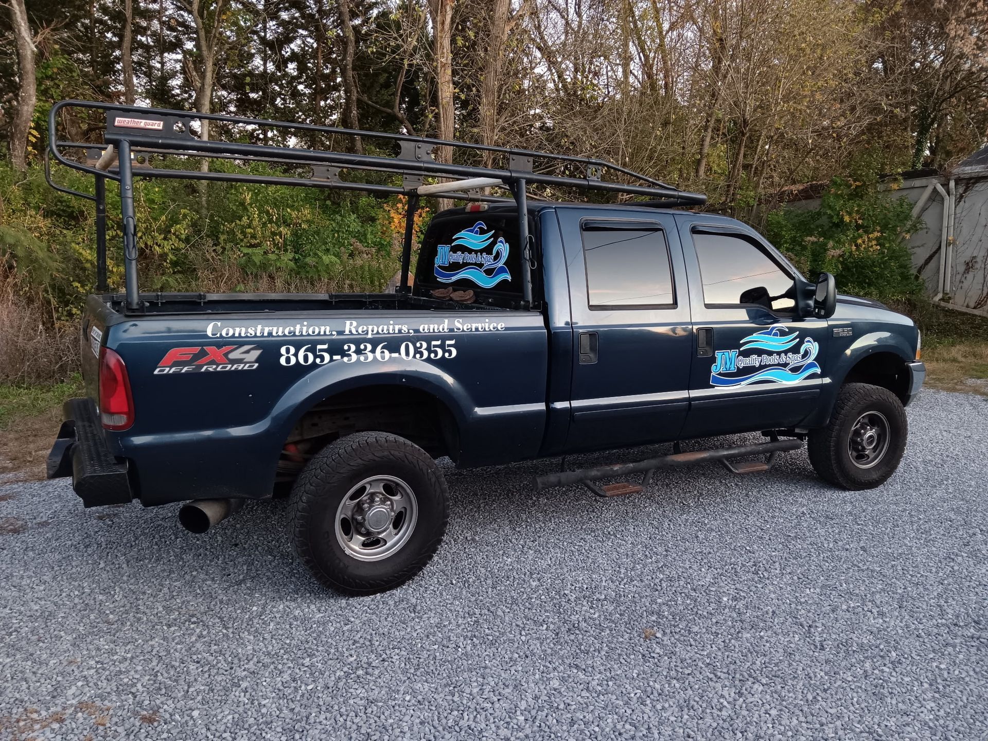A blue truck with a ladder rack on the back is parked on gravel.