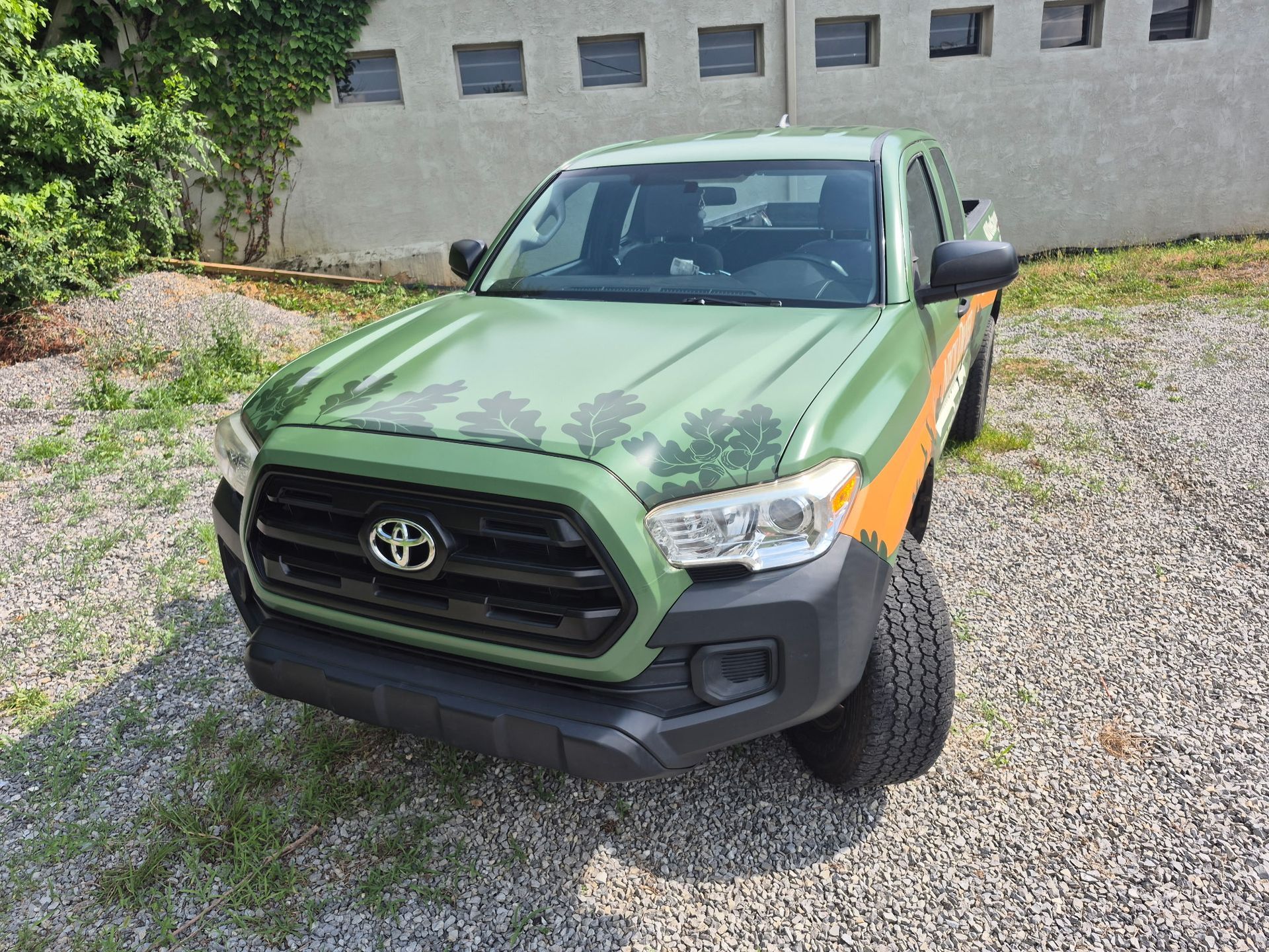 A green toyota tacoma truck is parked in a gravel lot in front of a building.