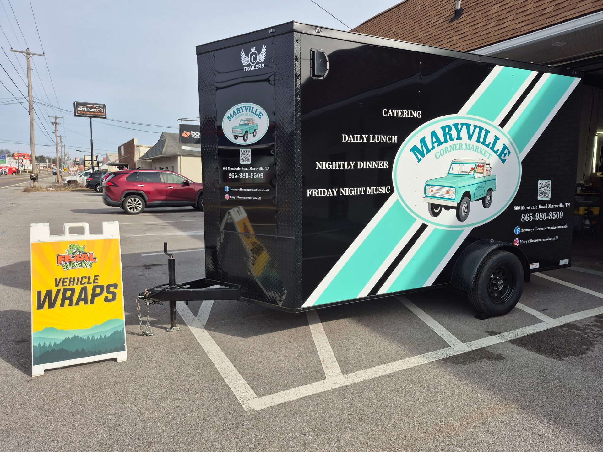 Food trailer in parking space, black exterior with teal accents, Maryville logo, advertising wraps.