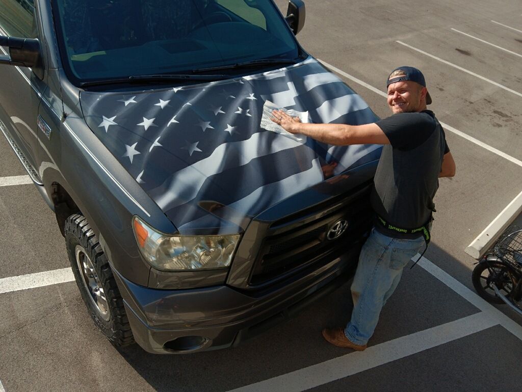 A man is standing next to a truck with an american flag on the hood.