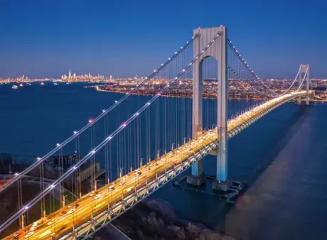 Verrazano-Narrows Bridge at dusk with city skyline in background, spanning blue water, illuminated lights.