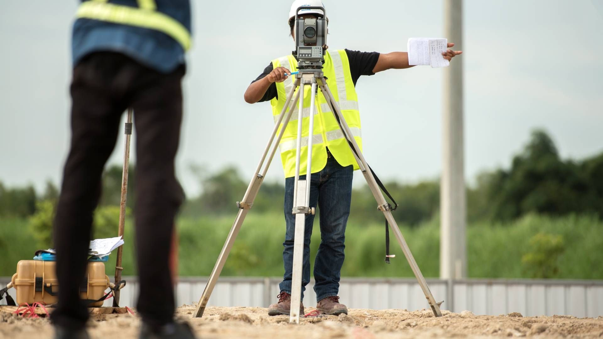 Surveyor in yellow vest uses a tripod-mounted instrument, pointing to construction site, with another worker nearby.