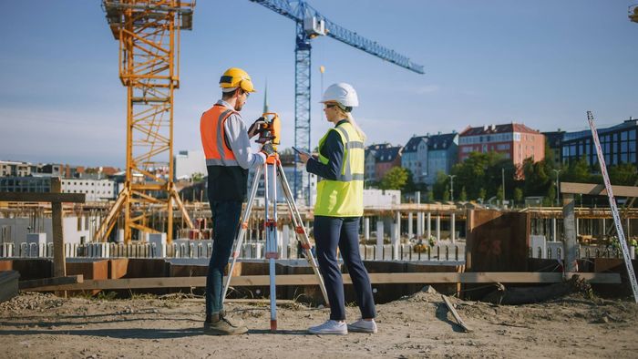 Two construction workers using surveying equipment at a building site with cranes and buildings in the background.