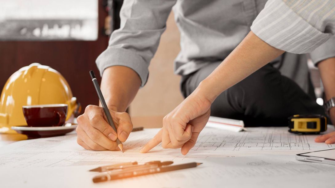 Two people pointing at and writing on blueprints with a yellow hard hat and coffee cup in the background.