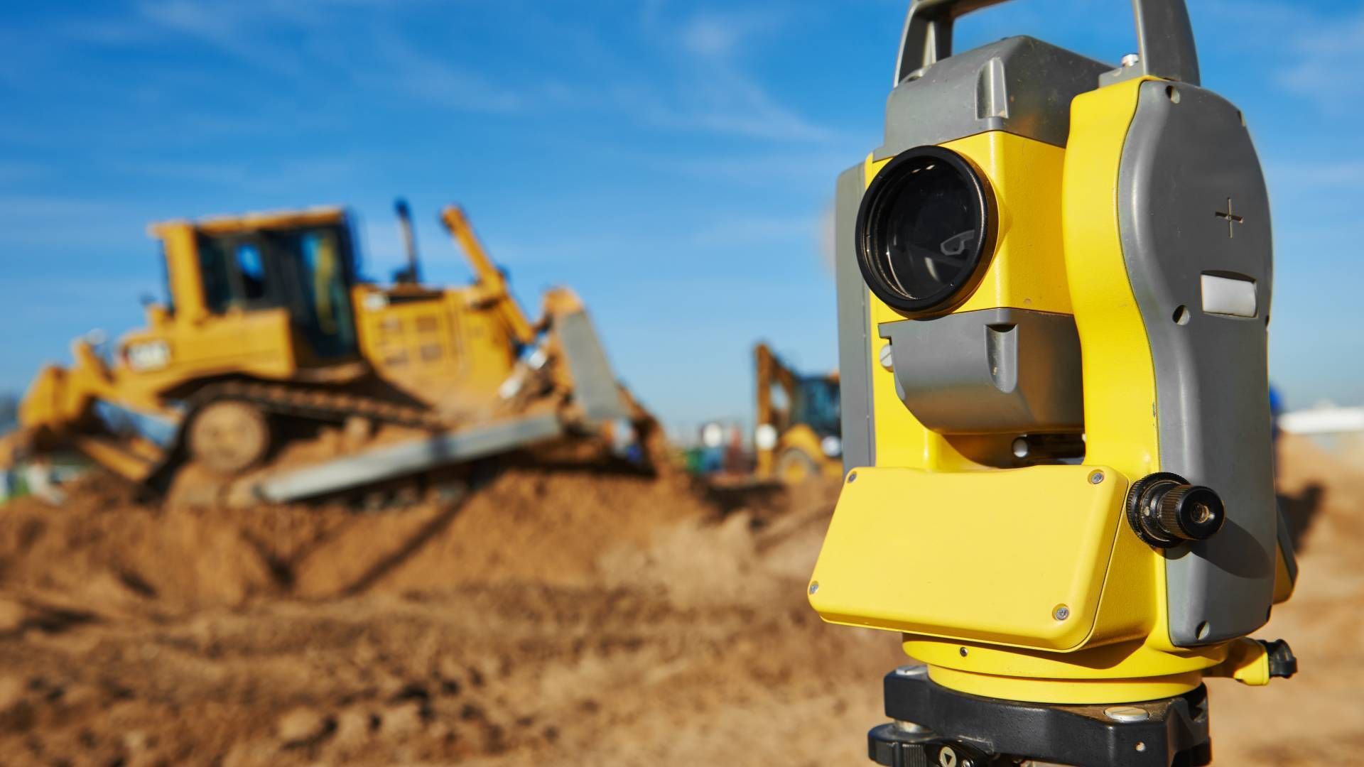 Yellow surveying instrument on tripod with construction equipment in the background.