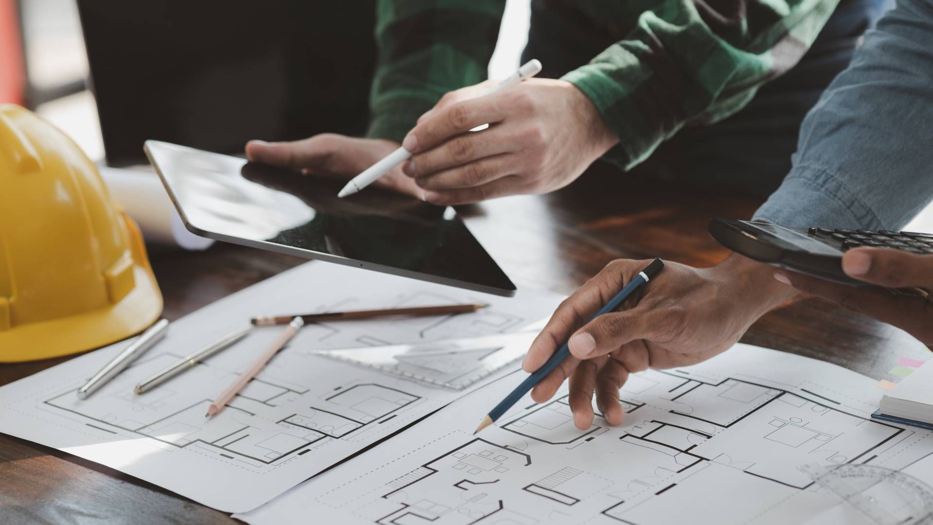 Architects reviewing blueprints, using a tablet, and a calculator at a table, with a yellow hard hat nearby.