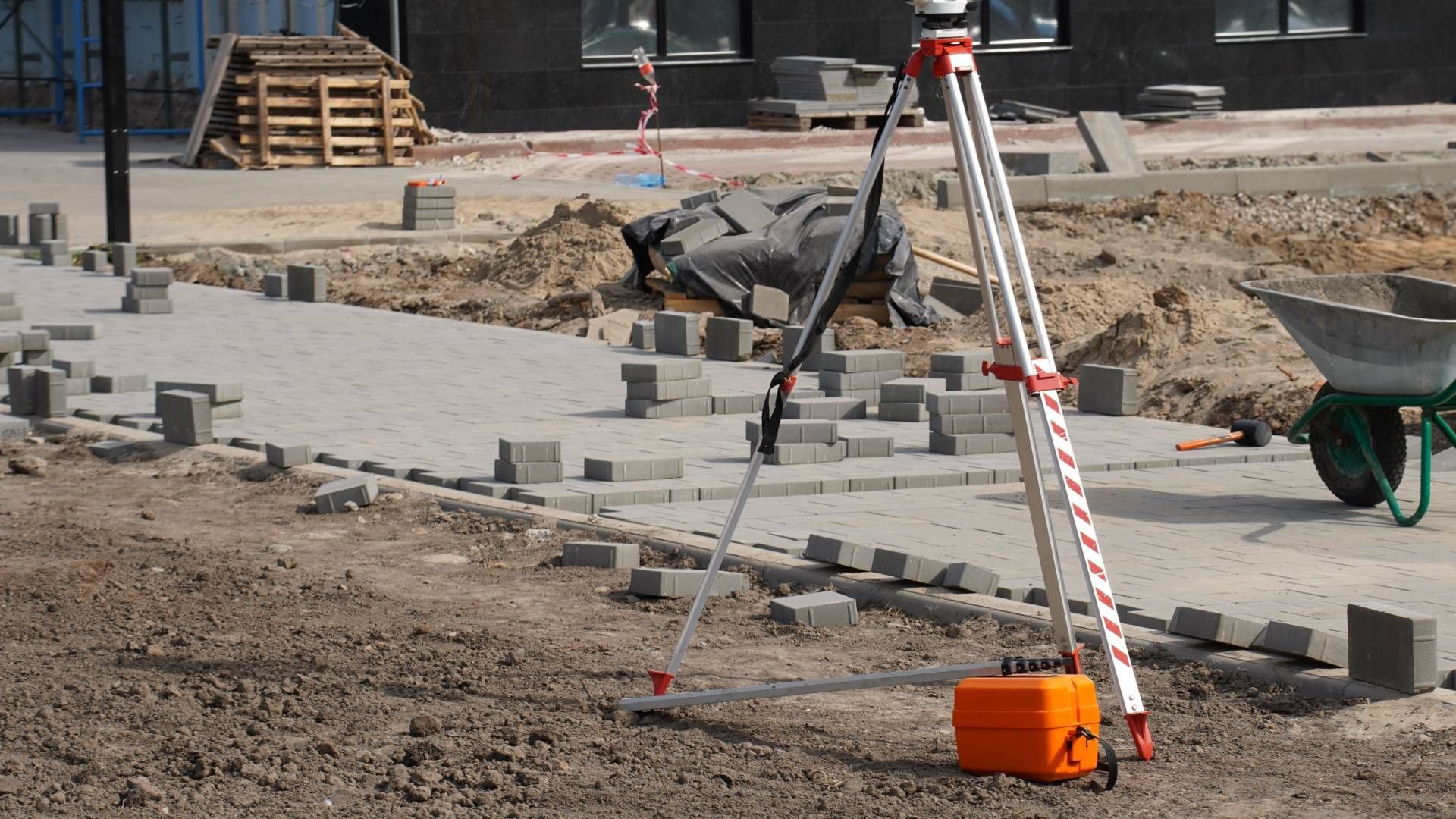 Construction site with paving blocks, surveying equipment, and a wheelbarrow.