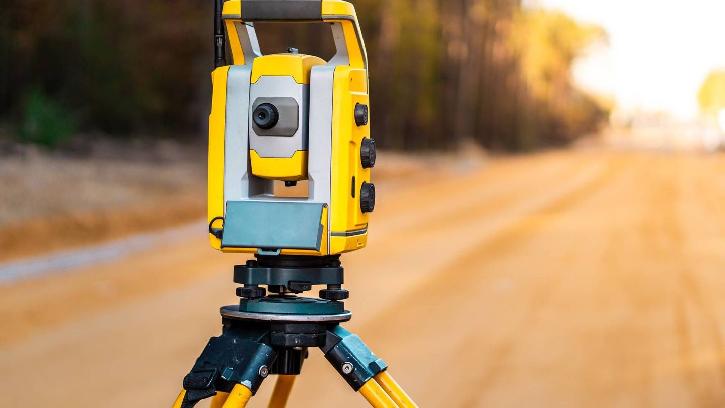 Yellow surveying instrument on a tripod, set on a dirt road.