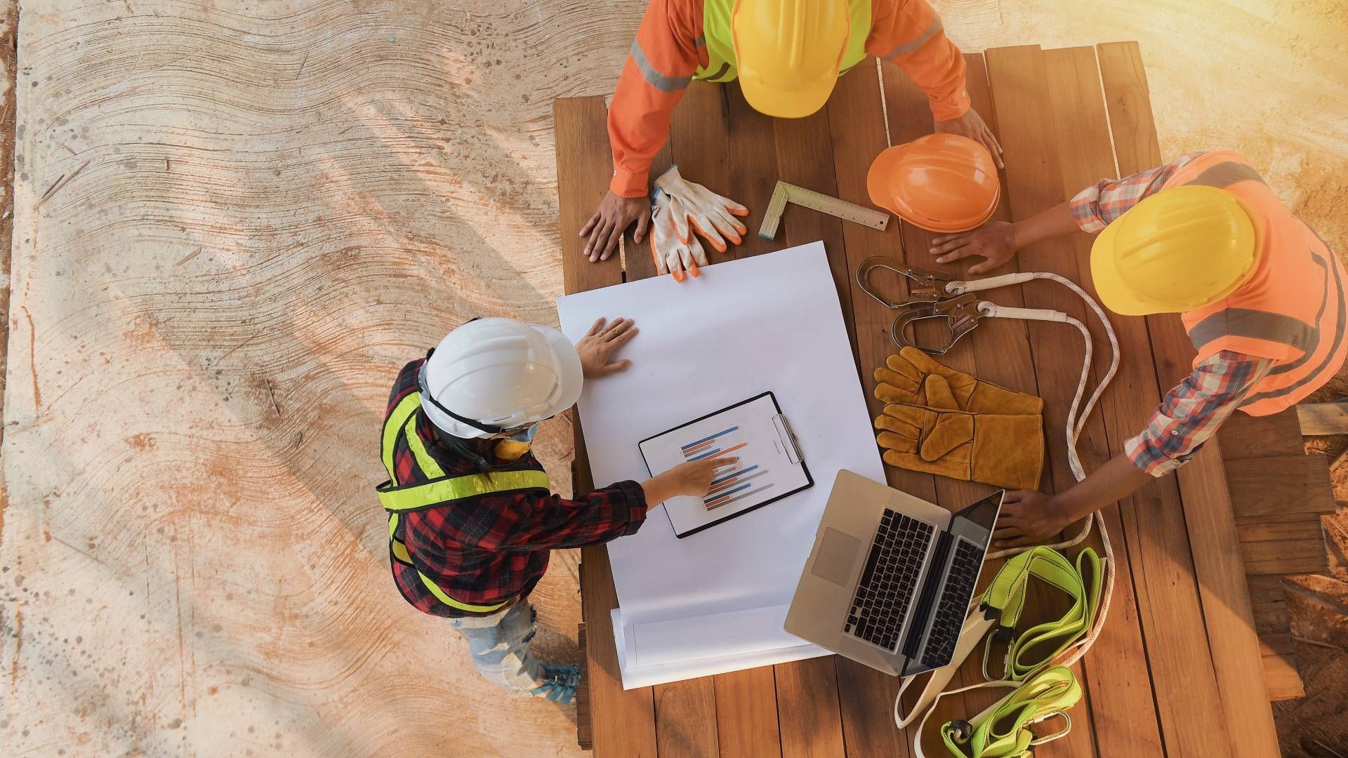 Construction workers in hard hats review blueprints around a table with a laptop.