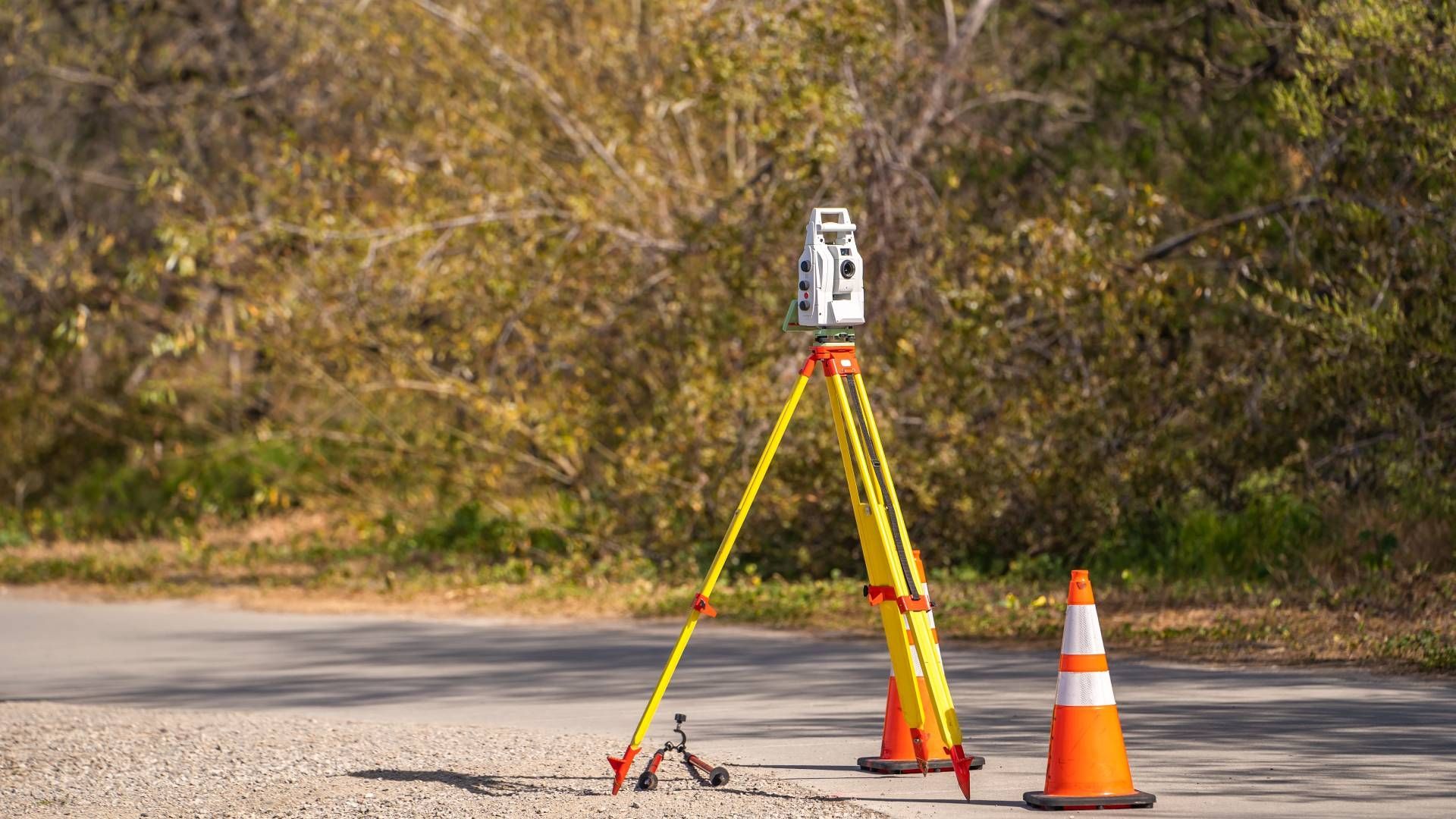 A surveying instrument on a tripod, orange safety cone, on a road with trees in the background.