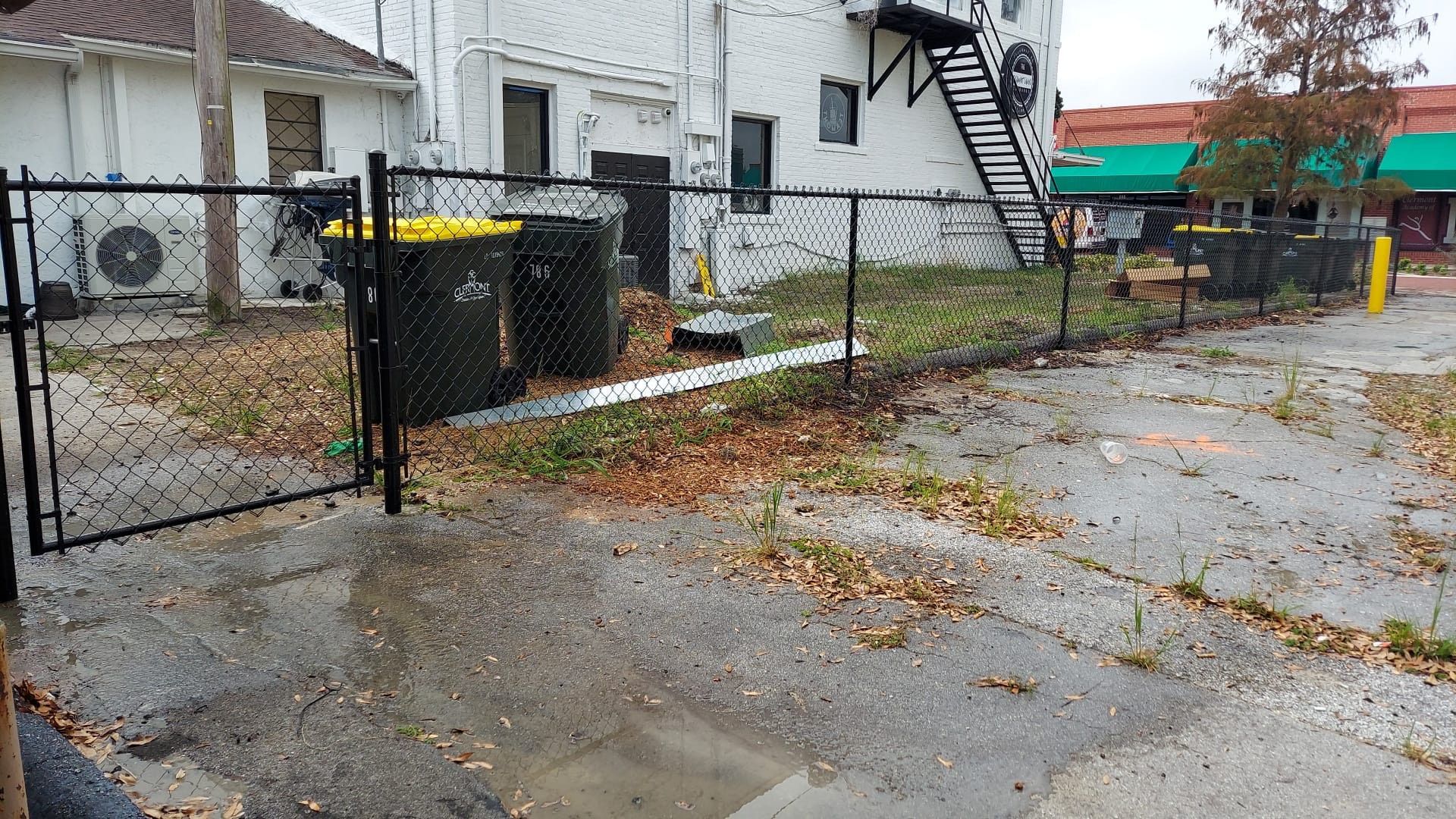 A chain link fence is surrounding a lot with trash cans and a building in the background.