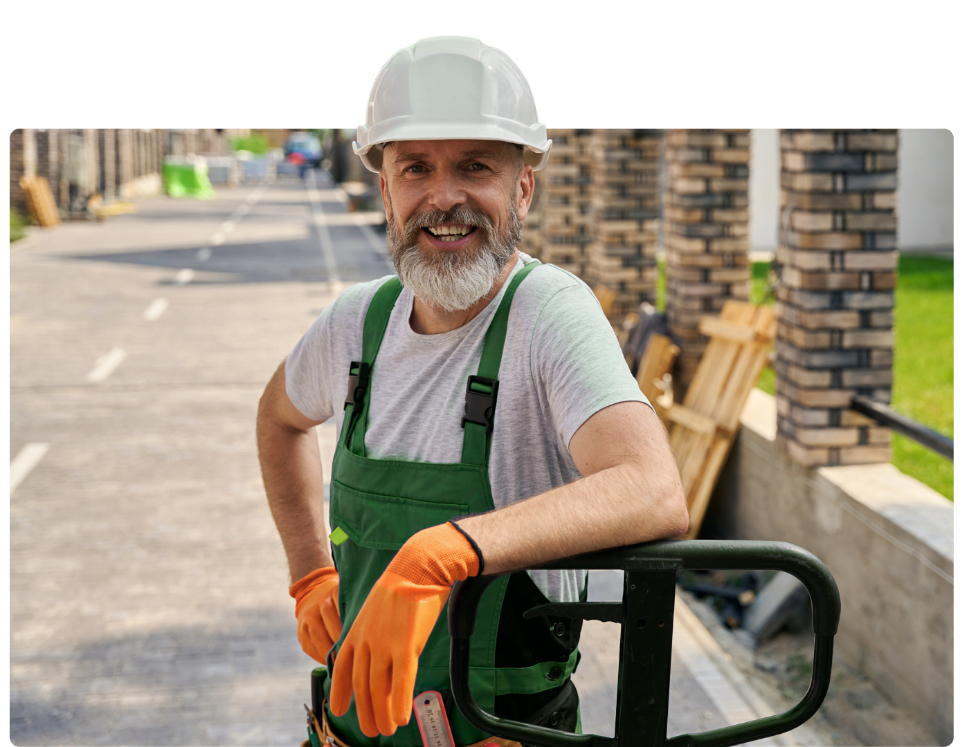 A man wearing a hard hat and overalls is leaning on a pallet truck.
