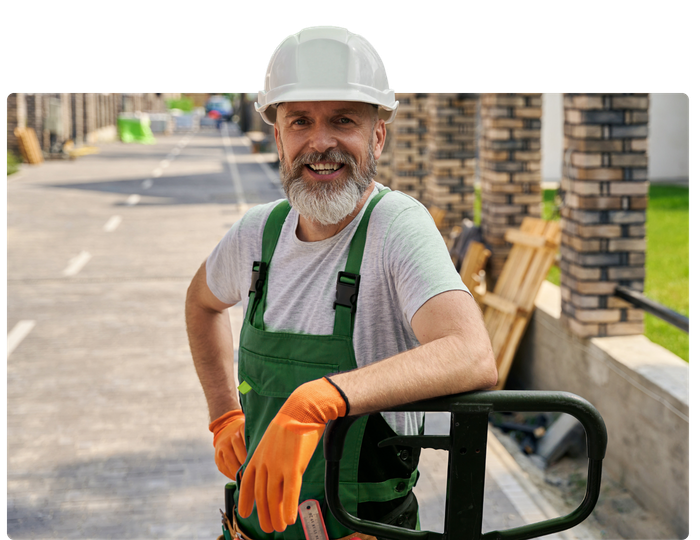 A man wearing a hard hat and overalls is leaning on a pallet truck.