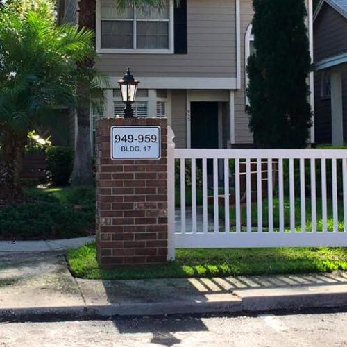 A white fence surrounds a brick pillar in front of a house.