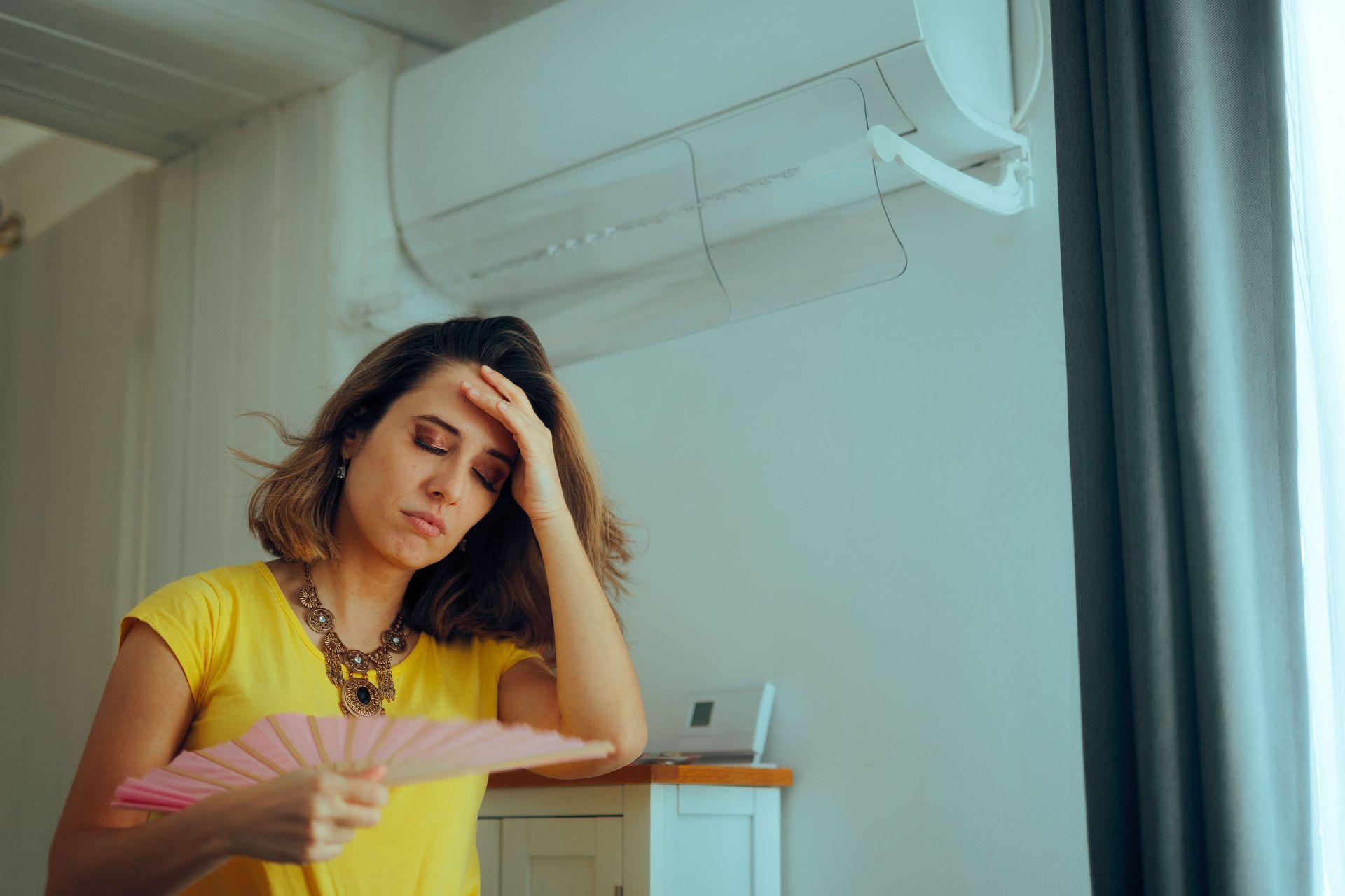 Woman looking overheated while fanning herself beneath a wall-mounted air conditioner.