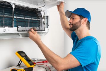 HVAC technician in blue shirt and cap servicing an air conditioner; tools on the floor.
