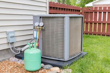 Air conditioner unit with gauges and a green refrigerant tank on a lawn near a wooden fence.