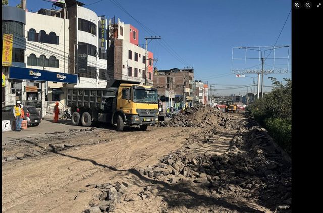 Calle urbana cubierta de barro con un camión volquete amarillo y superficie de la carretera dañada bajo un cielo azul claro.