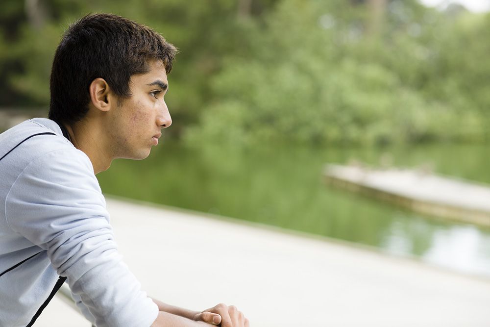 Young person looks pensively toward a lake, arm resting on a railing.