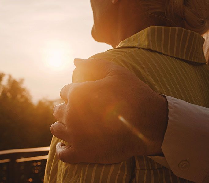 Hand on woman's shoulder as they look at a sunset; golden light illuminates them.