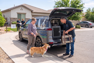 Two people unload supplies from a branded van while a dog stands nearby in a driveway